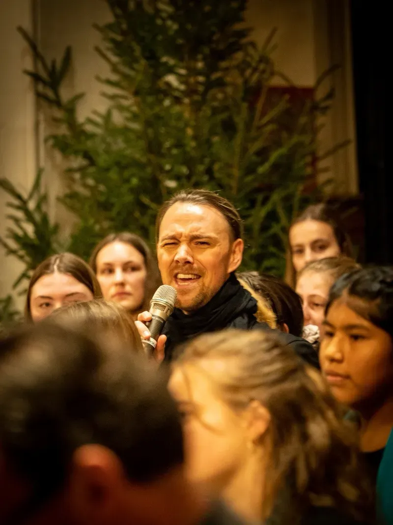 Maik singt mit einem Mikrofon inmitten der jungen Sängerinnen und Sänger der Singschule Sense.