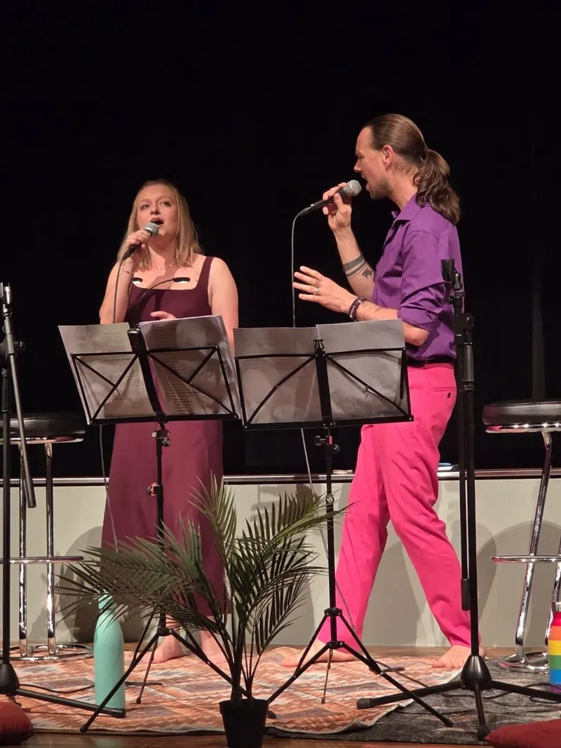 Maik und Chiara singen gemeinsam mit Mikrofonen und Notenständern auf einer Bühne.