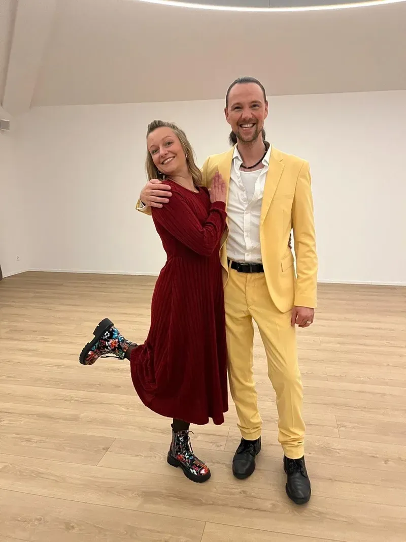 Chiara im roten Kleid und Maik im gelben Anzug stehen lächelnd nebeneinander.