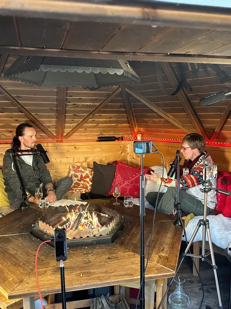Maik und Mirja sitzen mit Mikrofonen und Kameras am Feuer in einer hölzernen Grillhütte.