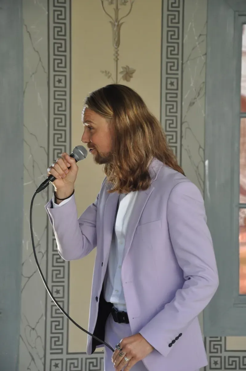 Maik singt mit einem Mikrofon im hellvioletten Anzug vor einer verzierten Wand.