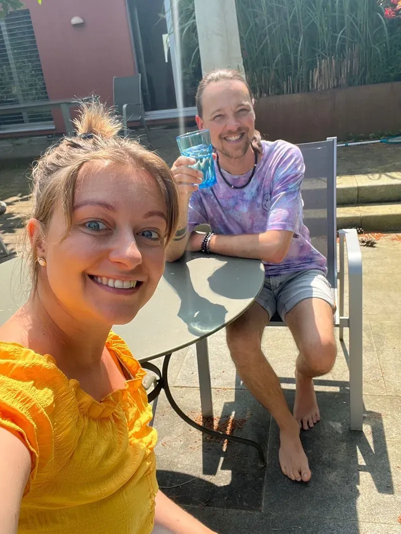 Chiara und Maik lächeln an einem Tisch auf einer sonnigen Terrasse in die Kamera.