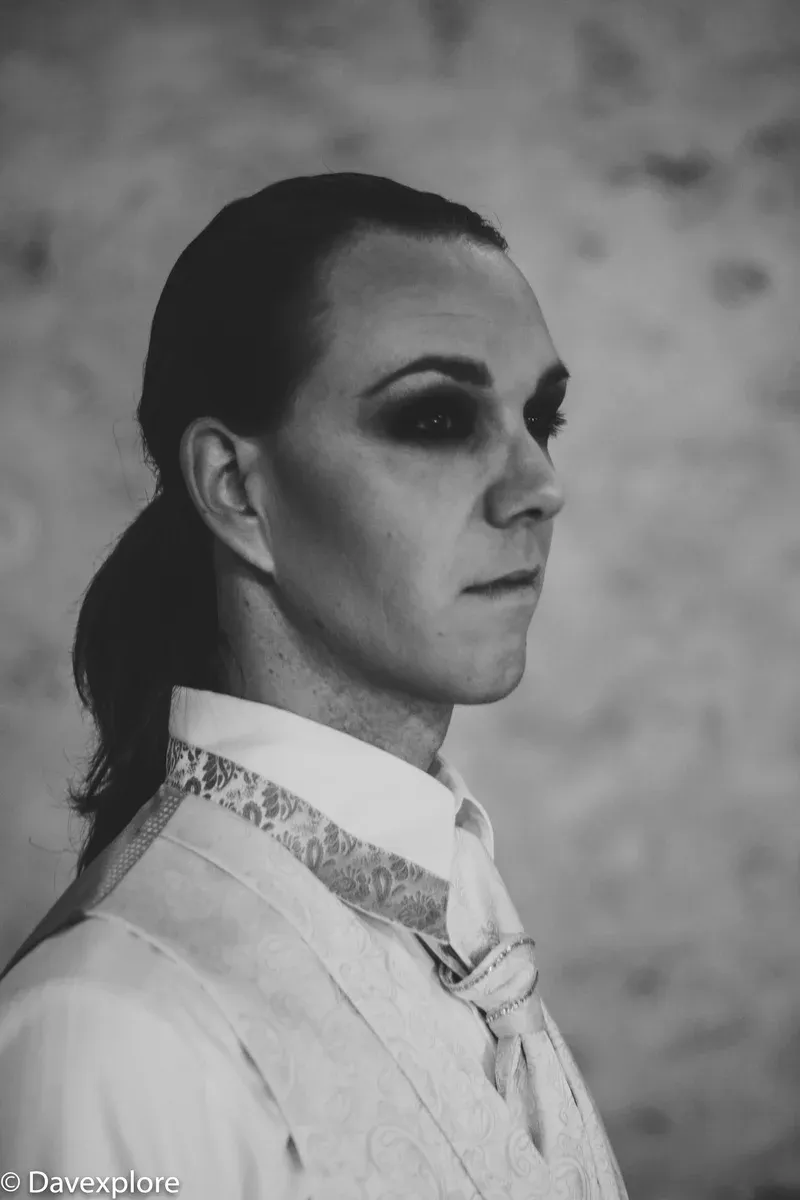 Schwarzweisses Porträt von Maik mit dunklem Augen-Make-up und heller gemusterter Weste.